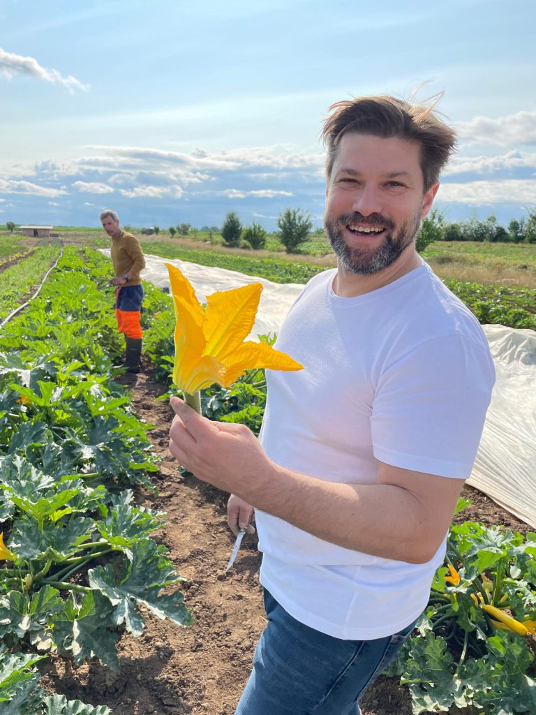 Portrait de Thierry Schwartz en plein cœur d'un champ de récolte, avec en arrière plan, un maraicher. Le Chef tient en main une fleur de courgette fraichement cueillie